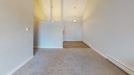 This single-floor residence offers a clean, functional layout with thoughtful finishes throughout. The galley-style kitchen is a standout feature, lined with crisp white shaker cabinetry, light countertops, and wood-look flooring underfoot. Black appliances — including a refrigerator, electric range, and dishwasher — provide a sleek contrast, while a pendant light fixture adds a decorative touch overhead. The kitchen connects naturally to the main living areas, which are defined by vaulted ceilings that lend an open, airy feel. Pendant lighting highlights the transition between the carpeted living space and the hard-surface dining or entry area. The living and bedroom spaces feature plush carpeting and soaring ceilings, with large sliding glass doors that open to a private balcony, filling the rooms with natural light and providing outdoor views. The bathroom is well-equipped with a full-size soaking tub and shower surround, a white vanity with light countertops, brushed nickel fixtures, and a mirrored medicine cabinet. A separate closet area adjacent to the bathroom offers convenient storage. The overall ambiance is bright, neutral, and move-in ready, with consistent finishes that create a cohesive and comfortable living environment throughout.