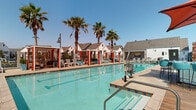 Located at 1575 Camino Altezza in Chula Vista, California, this single-level outdoor amenity space is designed to evoke a resort-like atmosphere within a residential community. The centerpiece is a generously sized swimming pool with crystal-clear turquoise water, framed by tall swaying palm trees that reinforce the Southern California ambiance. The pool deck is well-appointed with modern white lounge chairs and colorful accent pieces, offering ample space for sunbathing and relaxation.

Red-accented private cabanas line one side of the pool, providing shaded retreats with stylish seating for smaller gatherings. A dedicated outdoor kitchen area features granite countertops, a stainless steel sink, and a mounted flat-screen television set against a warm wood-panel accent wall, complemented by a living green wall that adds a natural, vibrant touch.

Covered lounge nooks are thoughtfully positioned around the perimeter, furnished with plush sectional seating in bold teal and red tones, framed artwork, and wall-mounted sconces. The surrounding architecture blends contemporary and traditional elements, with clean white facades, steel railings, and large windows. Manicured landscaping with succulents and shrubs enhances the outdoor environment, creating a polished, welcoming communal space ideal for year-round outdoor enjoyment.
