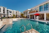 Located at 13525 Grayson Hi Cir in Dale City, Virginia, this expansive outdoor amenity area spans nearly 4,000 square feet of thoughtfully designed recreational space. At its centerpiece is a large, resort-style swimming pool with crystal-clear blue water, stone-finished water features, and stainless steel entry railings. An ADA-accessible pool lift ensures inclusive access for all users. The pool is framed by a generous poured-concrete sundeck furnished with rows of cushioned lounge chairs and striped market umbrellas, creating a comfortable environment for relaxation and outdoor leisure. A dedicated pergola area with additional seating provides a shaded social gathering space. Flanking one side of the pool is a modern two-story clubhouse building, distinguished by its contemporary mix of brick, board-and-batten siding, and large black-framed windows. The ground level features an open indoor-outdoor lounge and dining area, while a covered upper-level balcony overlooks the pool deck. Ornamental landscaping, iron perimeter fencing, and lush plantings add greenery and definition to the space. The entire area is set against a backdrop of multi-story residential buildings, tying the amenity space cohesively into the broader community. The open-sky setting ensures abundant natural light throughout the day.
