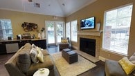 This single-floor space offers a warm and inviting atmosphere, thoughtfully designed for comfort and functionality. The main lounge area is the centerpiece of the space, highlighted by soaring vaulted ceilings with crown molding that lend an open, airy feel. Rich hardwood flooring runs throughout, anchoring the room with a sense of warmth and elegance.

A brick fireplace with a classic white mantle serves as a striking focal point, topped by a wall-mounted flat-screen television — a perfect blend of traditional charm and modern convenience. The seating arrangement includes a plush, generously sized sectional sofa accented with yellow and gray throw pillows, complemented by a pair of sleek ottoman cubes resting atop a soft, textured area rug.

Large windows dressed with white plantation shutters line the walls, flooding the space with natural light while offering views of the lush outdoor surroundings. French doors lead directly to an exterior courtyard area, further enhancing the connection between indoors and outdoors. A decorative sideboard along one wall adds additional storage and visual interest. The overall ambiance is relaxed yet polished, making this space well-suited for both casual gatherings and quiet relaxation.