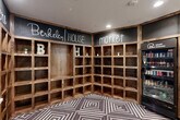 The Berkeley House Market is a thoughtfully designed self-serve retail space that blends rustic charm with modern convenience. The single-room layout makes efficient use of its compact footprint, offering a welcoming and functional environment for residents or guests.

Anchoring the space is a wrap-around shelving unit constructed from warm, natural wood with an open cubby design, ideal for displaying packaged goods and sundry items. Above the shelving, dark chalkboard panels display hand-lettered signage, including "Pay Here," "Berkeley House," and "Market," lending the space a casual, approachable aesthetic that balances style with practicality.

Two sleek black 24-hour smart vending machines are positioned along one wall, stocked with beverages, snacks, and packaged foods, ensuring round-the-clock accessibility. Recessed ceiling lighting illuminates the space evenly, highlighting the clean white walls and the eye-catching geometric diamond-patterned carpet in grey and white tones, which adds a dynamic visual element underfoot.

A large, vibrant painting of a cow on a blue background adorns one wall, injecting a playful artistic touch into the space. A keyed access door provides secure entry. Overall, the Berkeley House Market combines rustic warmth, modern vending technology, and distinctive design details to create an engaging and convenient amenity space.