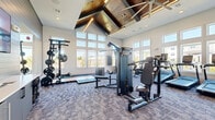This fitness center offers a generously sized, single-level workout space designed to accommodate a full range of exercise activities. The room's most striking architectural feature is its dramatic vaulted ceiling, accented with geometric wood paneling and integrated linear lighting that creates a warm, modern aesthetic. Rows of large windows line multiple walls, flooding the space with natural light and providing open views of the surrounding exterior.

The flooring throughout consists of patterned commercial-grade carpet tiles, providing cushioning underfoot. The walls feature a combination of neutral painted drywall and horizontal shiplap-style paneling in a muted gray tone, lending a contemporary, upscale character to the space.

Equipment is thoughtfully distributed throughout the room. The cardio zone includes multiple treadmills, elliptical machines, and a stationary cycling bike. A dedicated strength area features a multi-station cable machine, a power rack with weight plates, and a full dumbbell rack displayed alongside a large wall mirror that visually expands the space. Adjustable benches are positioned throughout for versatile use. Additional amenities include a small refrigeration unit near the entry and wall-mounted informational signage. Double glass doors provide easy access to the exterior.