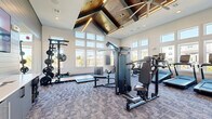 This fitness center offers a generously sized, single-level workout space designed to accommodate a full range of exercise activities. The room's most striking architectural feature is its dramatic vaulted ceiling, accented with geometric wood paneling and integrated linear lighting that creates a warm, modern aesthetic. Rows of large windows line multiple walls, flooding the space with natural light and providing open views of the surrounding exterior.

The flooring throughout consists of patterned commercial-grade carpet tiles, providing cushioning underfoot. The walls feature a combination of neutral painted drywall and horizontal shiplap-style paneling in a muted gray tone, lending a contemporary, upscale character to the space.

Equipment is thoughtfully distributed throughout the room. The cardio zone includes multiple treadmills, elliptical machines, and a stationary cycling bike. A dedicated strength area features a multi-station cable machine, a power rack with weight plates, and a full dumbbell rack displayed alongside a large wall mirror that visually expands the space. Adjustable benches are positioned throughout for versatile use. Additional amenities include a small refrigeration unit near the entry and wall-mounted informational signage. Double glass doors provide easy access to the exterior.