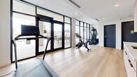 This contemporary fitness center is designed with both functionality and aesthetics in mind, offering a well-appointed workout space bathed in natural light. The room features light hardwood flooring throughout, crisp white ceilings with recessed lighting, and a built-in HVAC system to maintain a comfortable environment during workouts.

Large floor-to-ceiling windows and glass-paneled doors line one wall, providing abundant natural light and views of an adjacent outdoor terrace, which is furnished for additional relaxation or cool-down space. The open layout accommodates a variety of cardio and strength equipment, including treadmills, a Peloton spin bike, and a multi-station cable machine, all arranged to maximize usable floor space.

Along one wall, a built-in cabinetry unit with a light wood finish and a white countertop with a sink serves as a convenient wet bar or hydration station. A textured concrete accent wall adds an industrial contrast to the otherwise clean, minimalist interior. Two separate doors — one marked as an exit and another appearing to lead to restroom facilities — provide practical access points. The overall design blends modern architecture with a welcoming, professional atmosphere well-suited for focused fitness activity.