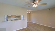 This single-floor residence offers a welcoming open-concept design that seamlessly connects the living area and kitchen. The living room is finished with wall-to-wall carpeting in a warm neutral tone, creating a comfortable and cohesive atmosphere throughout the space. A ceiling fan with an integrated light fixture anchors the main living area, providing both functionality and a casual aesthetic. An elegant small chandelier adds a touch of refinement to the adjoining room, enhancing the overall ambiance. The kitchen is accessible via a raised breakfast bar counter, which serves as a natural divider between the cooking and living spaces while maintaining the open feel. From the living area, a pass-through window offers a clear sightline into the kitchen, where a range and cabinetry are visible. Neutral-toned walls throughout the space lend a clean, versatile backdrop suited to a variety of décor styles. White baseboards and trim provide crisp contrast against the soft carpeting and walls. The layout flows naturally, making the space feel airy and connected, with practical features thoughtfully integrated into the design.