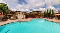 Located at 12302 S Yukon Ave in Glenpool, Oklahoma, this outdoor amenity space centers around a generously sized freeform swimming pool with a smooth concrete and stone surround. The pool's organic shape adds a resort-like character to the area, complemented by a separate hot tub tucked along one edge. A charming wood pergola provides a shaded gathering spot, furnished with red Adirondack chairs — perfect for relaxing out of direct sun. Surrounding the pool, an array of reclining lounge chairs and umbrella-shaded tables offer ample space for sunbathing and outdoor dining. A dedicated pool house structure sits on the premises, featuring siding, a pitched roof, and a private entrance, likely housing restrooms or storage facilities. The entire area is enclosed by a wrought-iron fence, ensuring privacy and security. Lush landscaping with ornamental grasses, flowering shrubs, cypress trees, and mature shade trees frames the space with vibrant greenery. The backdrop of the multi-story residential building visible beyond the fence adds architectural context, while the open sky above allows for abundant natural light throughout the day.