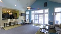 This single-floor space makes a strong first impression with its dramatic vaulted ceilings that lend an airy, expansive feel to the interior. The open-concept layout is anchored by a wall of large French doors with transom windows above, flooding the space with abundant natural light and offering views to the outdoors. A ceiling fan with a decorative light fixture hangs centrally, complementing the recessed lighting throughout.

The interior is divided into functional zones that flow seamlessly into one another. A casual lounge area features plush seating arranged around a glass-top coffee table atop a textured area rug, ideal for relaxed gatherings. Adjacent to it, a dining area is furnished with a round table and a mix of upholstered chairs, set beneath a wall-mounted flat-screen television. A bar-height counter with tall white chairs creates a third seating option, adding to the social versatility of the space.

Color plays a key role in defining the ambiance — a bold teal accent wall near the French doors contrasts with warm sage and neutral tones elsewhere, creating a vibrant yet cohesive atmosphere. The carpeted flooring, layered with area rugs, adds warmth and comfort underfoot. Overall, the space is thoughtfully designed to encourage community interaction in a welcoming and well-lit environment.
