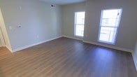 This single-floor space offers a clean, move-in-ready interior designed with simplicity and functionality in mind. The main living area is immediately inviting, featuring smooth hardwood-style plank flooring in a warm, natural tone that extends throughout the space, creating a cohesive and polished look underfoot. The walls are finished in a soft, neutral beige that complements the flooring and provides a versatile backdrop for any décor style. Natural light is a defining feature of this space, with multiple windows along the exterior walls allowing generous daylight to flow in and illuminate the interior throughout the day. White baseboards frame the perimeter of the room, adding a crisp, finished detail that ties together the overall aesthetic. A ceiling-mounted HVAC vent ensures year-round climate comfort, while electrical outlets are thoughtfully positioned along the walls for everyday convenience. The open layout of the main area provides ample room to configure a variety of living arrangements, making it a flexible and functional space. The overall ambiance is bright, clean, and understated — well-suited for those who appreciate a fresh, neutral canvas ready to be personalized.