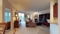 This single-floor residence offers a thoughtfully designed living area that serves as the central hub of the space. The living room is generously proportioned, featuring plush carpeting and warm, neutral-toned walls that create an inviting atmosphere. A standout feature is the tile-surround fireplace, which anchors one wall and adds both warmth and architectural interest to the room.

Built-in wood shelving units flank the entertainment area, providing ample display and storage space while contributing a rich, cohesive aesthetic. Large sliding glass doors flood the living space with natural light and open onto a private balcony overlooking lush, green surroundings — a notable feature that enhances the overall sense of openness.

The layout connects seamlessly to an adjacent area, visible through an open passageway, where recessed lighting and a white entry door suggest a well-lit entryway. A round wooden accent table with a decorative plant centerpiece and coordinating furniture in warm earth tones complete the space, offering a comfortable and functional environment throughout.