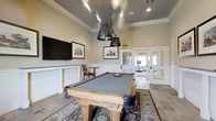 The billiards room is a thoughtfully designed recreational space that combines classic styling with functional amenities. At its center sits a substantial wood-framed pool table with a gray felt surface, positioned atop a decorative area rug and flanked by elegant pendant lights with industrial-style metal shades that hang from a tray ceiling, adding both task lighting and visual character to the room.

The elevated tray ceiling enhances the sense of height and architectural detail, while crown molding along the upper walls contributes a refined, traditional aesthetic. The walls are finished in a warm neutral tone and adorned with framed nature-themed artwork, complementing the classic decor. A wall-mounted flat-screen television is installed to one side, offering entertainment beyond the game table, and a small side table with seating provides a comfortable spot for spectators.

The flooring transitions between tile and an area rug beneath the pool table, adding texture and warmth. French doors at the rear of the room open to an adjacent fitness area, allowing easy access between recreational spaces. The overall ambiance is inviting and polished, ideal for leisure and social gatherings.