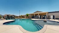 Located in El Paso, TX, this two-story community recreation facility centers around a standout resort-style pool that serves as the focal point of the outdoor amenity space. The freeform pool features a striking rock waterfall feature, a decorative fountain, and defined entry points with steel handrails, all finished with a classic blue tile border. The expansive surrounding pool deck provides ample room for lounging, with multiple chaise lounge chairs and patio seating areas positioned throughout the space.

A covered clubhouse structure anchors one side of the pool area, featuring a gabled roofline supported by decorative columns and an inviting covered porch. The building's exterior reflects a warm earth-tone stucco finish complemented by stone-accented pillar details that are carried throughout the perimeter fencing, lending a cohesive Southwestern aesthetic to the entire complex.

The grounds are enclosed by a wrought iron and stone-pillar security fence, with lush desert-adapted landscaping including palm trees, ornamental shrubs, and decorative boulders that enhance the resort ambiance. Additional outdoor seating with tables and chairs provides shaded gathering areas along the perimeter. Under El Paso's expansive blue skies, the space offers a bright, open atmosphere well-suited to the warm desert climate of West Texas.