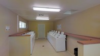 This single-floor communal laundry facility is designed with practicality and efficiency in mind. The room is well-lit by overhead fluorescent fixtures that illuminate the entire space evenly, creating a bright and functional environment. A row of top-loading washing machines lines the center of the room, paired with a matching set of dryers, providing ample capacity to serve multiple users simultaneously. Along the perimeter, generous counter space with a contrasting trim offers a convenient surface for sorting and folding laundry. The walls are painted in a warm neutral tone, contributing to a clean and welcoming atmosphere. Ventilation is provided through ceiling vents, ensuring proper airflow throughout the space. A single door at the far end of the room provides access, and a window along one wall allows for a touch of natural light to supplement the overhead lighting. The flooring is a light-toned, easy-to-clean surface suited for a high-traffic utility space. Informational notices are posted on the walls, keeping users informed of guidelines and instructions. A waste bin is conveniently positioned near the entrance for easy disposal. Overall, this laundry facility is thoughtfully arranged to maximize usability and comfort for its users.