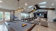 Located at 9005 Oak Village Blvd in Lewis Center, Ohio, this single-story community clubhouse offers a well-appointed gathering space designed to accommodate a variety of activities. The centerpiece of the facility is a generously sized game and lounge area highlighted by dramatic vaulted ceilings with exposed dark wood beams and multiple skylights that flood the room with natural light. This space features a full-size pool table with a stylish industrial pendant light fixture, a shuffleboard table, oversized Jenga, and comfortable seating including a tufted leather sofa and plush sectionals — ideal for socializing and recreation. A wall-mounted flat-screen TV and a textured accent wall in deep charcoal add a modern, polished aesthetic throughout. The kitchenette area is outfitted with rich forest green cabinetry, gold hardware, a geometric tile backsplash, a sink, and a beverage refrigerator. Adjacent to the main lounge, a dedicated business center provides computer workstations, a round conference table with leather chairs, and large windows overlooking the surrounding neighborhood. A separate meeting nook features a wall-mounted display and upholstered seating. Warm wood-look flooring runs throughout, tying the space together with a cohesive, contemporary feel.