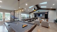 Located at 9005 Oak Village Blvd in Lewis Center, Ohio, this single-story community clubhouse offers a well-appointed gathering space designed to accommodate a variety of activities. The centerpiece of the facility is a generously sized game and lounge area highlighted by dramatic vaulted ceilings with exposed dark wood beams and multiple skylights that flood the room with natural light. This space features a full-size pool table with a stylish industrial pendant light fixture, a shuffleboard table, oversized Jenga, and comfortable seating including a tufted leather sofa and plush sectionals — ideal for socializing and recreation. A wall-mounted flat-screen TV and a textured accent wall in deep charcoal add a modern, polished aesthetic throughout. The kitchenette area is outfitted with rich forest green cabinetry, gold hardware, a geometric tile backsplash, a sink, and a beverage refrigerator. Adjacent to the main lounge, a dedicated business center provides computer workstations, a round conference table with leather chairs, and large windows overlooking the surrounding neighborhood. A separate meeting nook features a wall-mounted display and upholstered seating. Warm wood-look flooring runs throughout, tying the space together with a cohesive, contemporary feel.