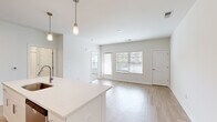 Located at 44000 Owl Wood Lane in Summerville, SC, this one-bedroom, one-bathroom residence offers a thoughtfully designed single-floor layout with clean, contemporary finishes throughout. The open-concept living and kitchen area is bright and airy, featuring light hardwood-style flooring, white walls, and large windows fitted with blinds that allow ample natural light to fill the space. The kitchen is well-appointed with white shaker-style cabinetry extending to the ceiling, a white quartz countertop island with pendant lighting overhead, and a full suite of stainless steel appliances including a refrigerator, electric range, microwave, and dishwasher. A white subway tile backsplash adds a polished touch. The living room offers a comfortable, open feel with additional windows providing outdoor views. The bedroom features soft carpet flooring, a ceiling fan, and a window with natural light. The bathroom is finished in white with a single-sink vanity, white cabinetry, wood-look tile flooring, and a full tub and shower combination with white subway tile surround. A dedicated laundry room houses a full-size washer and dryer with overhead wire shelving for added storage. A closet completes the layout, offering practical everyday convenience.