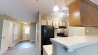 This single-floor residence offers a practical and open layout designed for comfortable everyday living. The kitchen serves as a central hub, featuring a mix of white and warm wood-toned upper cabinetry that provides ample storage. A black refrigerator and black gas range anchor the cooking space, complemented by an under-cabinet range hood. A white breakfast bar counter extends outward, creating a casual dining or prep area that opens the kitchen toward the adjoining living space. Overhead, a pendant light and a track lighting system ensure the kitchen is well-lit throughout the day. The flooring transitions from hard surface in the kitchen area to carpet in the living space, adding warmth underfoot. A hallway connects the kitchen and living areas to the front entryway, where a windowed door allows natural light to filter into the interior, brightening the corridor. The neutral wall tones throughout contribute to a clean, cohesive aesthetic, while the open sightlines between rooms create a sense of connected, functional space suited to daily routines.