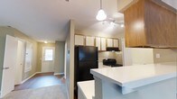This single-floor residence offers a practical and open layout designed for comfortable everyday living. The kitchen serves as a central hub, featuring a mix of white and warm wood-toned upper cabinetry that provides ample storage. A black refrigerator and black gas range anchor the cooking space, complemented by an under-cabinet range hood. A white breakfast bar counter extends outward, creating a casual dining or prep area that opens the kitchen toward the adjoining living space. Overhead, a pendant light and a track lighting system ensure the kitchen is well-lit throughout the day. The flooring transitions from hard surface in the kitchen area to carpet in the living space, adding warmth underfoot. A hallway connects the kitchen and living areas to the front entryway, where a windowed door allows natural light to filter into the interior, brightening the corridor. The neutral wall tones throughout contribute to a clean, cohesive aesthetic, while the open sightlines between rooms create a sense of connected, functional space suited to daily routines.