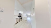 This thoughtfully designed 2-bedroom, 1-bathroom unit offers a clean, modern aesthetic across a single floor. The open-concept kitchen is a standout feature, outfitted with sleek, high-gloss white cabinetry extending to the ceiling, stainless steel appliances including a range, over-the-range microwave, dishwasher, and refrigerator, as well as a white quartz countertop and a peninsula that provides additional workspace and visual separation from the hallway. Recessed lighting and a window at the far end bring natural light into the kitchen and adjoining areas.

Light-toned hardwood-style flooring flows consistently throughout the unit, complementing the bright white walls and creating a cohesive, airy atmosphere. Both bedrooms are well-proportioned, with the primary bedroom offering generous space. One bedroom features a large window with an outdoor treetop view and a roller shade, while the other includes a double-door closet. The bathroom is finished with a clean white tile surround enclosing a soaker tub and shower combination with modern chrome fixtures. A connecting hallway ties all rooms together efficiently, contributing to a practical and functional layout overall.