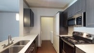 This single-floor unit showcases a contemporary design with consistent finishes throughout. The kitchen is a standout feature, outfitted with dark espresso-toned cabinetry, white countertops, and a full suite of stainless steel appliances, including a gas range, built-in microwave, and a side-by-side refrigerator with an in-door water and ice dispenser. A distinctive X-pattern wine rack tops the cabinetry, adding a stylish accent. The galley-style layout maximizes efficiency, with a stainless steel sink on one side and ample counter and storage space. Wood-look flooring runs through the kitchen and extends into the living area, where a ceiling fan with light, crown molding, and a sliding glass door with vertical blinds create a bright, comfortable atmosphere. The bedroom features plush carpeting, a large window fitted with blinds for natural light control, and a ceiling light fixture. The bathroom is clean and functional, offering a soaking tub with shower, a curved curtain rod, dark cabinetry for storage, and wood-look flooring. A dedicated in-unit laundry room houses a full-size top-loading washer and dryer alongside an overhead shelf, providing convenient and practical storage.