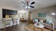 This single-floor residence offers a thoughtfully designed open-concept layout that seamlessly connects the living, dining, and kitchen areas. Dark hardwood-style flooring runs throughout the main living spaces, providing a warm and cohesive aesthetic. The living room is bright and inviting, featuring a ceiling fan, a large sliding glass door that floods the space with natural light, and direct access to a private patio overlooking lush greenery. The dining area accommodates a round wooden table and is accented by a decorative pendant light and an ornate framed mirror. The galley-style kitchen is well-equipped with dark cabinetry, light countertops, a double sink, and black appliances including a range, microwave, and refrigerator, with a breakfast bar providing additional seating. The primary bedroom is spacious and bright, with a ceiling fan, large windows, and dark hardwood-style flooring. Its en-suite bathroom features dark wood cabinetry, a wide vanity mirror, and a shower-tub combination. The second bedroom mirrors the comfortable feel of the primary, also featuring a ceiling fan and generous natural light. Its adjoining bathroom offers a tub-shower combination with neutral tones. The space maintains a consistent coastal-inspired design theme throughout.
