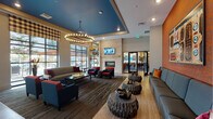 Located at 11001 W 15th Pl in Lakewood, Colorado, this single-floor community space is thoughtfully designed to offer a welcoming and versatile environment for its residents. The main lounge area immediately draws attention with its soaring ceilings, a bold blue tray ceiling detail, and dramatic ring chandeliers that cast warm, inviting light throughout. A stone-clad fireplace serves as a striking focal point, flanked by comfortable seating arrangements featuring navy and red upholstered sofas and chairs. Distinctive carved log side tables add a rustic, nature-inspired accent to the contemporary design. Large floor-to-ceiling windows with plaid Roman shades flood the space with natural light while offering views of an outdoor patio area. An accent wall finished in a rich copper-orange tone is adorned with a large-scale artwork, adding to the vibrant and eclectic character of the lounge. A grid of white-framed mirrors on an adjacent wall enhances depth and brightness. The dining area accommodates generous seating beneath matching statement chandeliers, supported by sturdy columns and complemented by a raised platform floor. The kitchen features sleek grey cabinetry, crisp white countertops, stainless steel appliances, and a wall-mounted television, creating a fully functional and stylish culinary space ideal for gatherings and events.