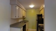 This single-floor space includes a galley-style kitchen designed for efficient use of space. The kitchen features white raised-panel cabinetry lining both sides of the corridor layout, providing generous storage. A stainless steel double sink is set into a gray laminate countertop along one wall, accompanied by a black dishwasher beneath. On the opposite side, a black electric range with coil burners sits adjacent to a black refrigerator, completing the appliance lineup. Additional upper cabinetry at the far end of the kitchen offers further storage capacity. The walls are painted a light yellow-green, adding a subtle warmth to the space. Tile flooring runs throughout the kitchen, offering durability and ease of maintenance. Overhead, a flush-mount ceiling light provides consistent illumination across the workspace. The overall layout is practical and straightforward, making efficient use of the available footprint while keeping all key kitchen functions within easy reach.