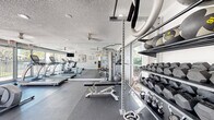Located at 2554 Oak Trail S in Clearwater, Florida, this single-floor fitness center offers a comprehensive workout environment designed to support a wide range of exercise routines. The space is outfitted with durable rubber flooring throughout, providing both safety and comfort during training sessions.

The cardio zone features multiple treadmills, elliptical trainers, and a recumbent bike, all positioned in front of expansive floor-to-ceiling windows that look out onto a resort-style pool area framed by palm trees — a bright, motivating backdrop for any workout. Large mirrors line one wall, enhancing the sense of space and allowing users to monitor their form.

The strength training area is equally well-appointed, with a multi-function cable machine, an adjustable weight bench, stability balls, and a full rack of dumbbells and kettlebells in varying weights. Ceiling fans are distributed throughout to maintain comfortable airflow, complementing the natural light that pours in through the large windows.

A textured ceiling and recessed lighting complete the clean, functional aesthetic. An adjacent room, visible through glass doors, appears to serve as an office or additional amenity space. The overall design is bright, open, and purpose-built for effective fitness training.
