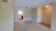 This single-floor residence offers a straightforward and functional layout designed for comfortable everyday living. The main living area is carpeted and finished with soft, neutral-toned walls that create a calm and understated atmosphere. A flush-mount ceiling light provides overhead illumination, while white baseboards frame the perimeter of the room cleanly. The entry door is conveniently positioned along one wall, offering direct access to the living space. Adjacent to the main area, the kitchen is accessible through an open doorway and features light wood-toned cabinetry, a sink positioned beneath a window that allows natural light to filter in, and a dark appliance — likely a dishwasher — alongside the cabinetry. The kitchen flooring transitions to a hard surface, providing a practical contrast to the carpeted living area. The overall design is clean and simple, with a neutral color palette throughout that lends itself well to personalization. The space is well-suited for those seeking a low-maintenance, move-in ready environment with a cohesive and uncluttered aesthetic.