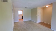 This single-floor residence offers a straightforward and functional layout designed for comfortable everyday living. The main living area is carpeted and finished with soft, neutral-toned walls that create a calm and understated atmosphere. A flush-mount ceiling light provides overhead illumination, while white baseboards frame the perimeter of the room cleanly. The entry door is conveniently positioned along one wall, offering direct access to the living space. Adjacent to the main area, the kitchen is accessible through an open doorway and features light wood-toned cabinetry, a sink positioned beneath a window that allows natural light to filter in, and a dark appliance — likely a dishwasher — alongside the cabinetry. The kitchen flooring transitions to a hard surface, providing a practical contrast to the carpeted living area. The overall design is clean and simple, with a neutral color palette throughout that lends itself well to personalization. The space is well-suited for those seeking a low-maintenance, move-in ready environment with a cohesive and uncluttered aesthetic.