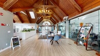 Located at 110 Mountainhigh Dr in Nashville, TN, this single-floor community facility is defined by its dramatic architectural character and versatile layout. The most commanding feature is the soaring vaulted ceiling clad in warm, rich wood planking, supported by exposed heavy timber beams that run the length of the space. Skylights and large arched windows — including a distinctive arched transom above the main entry doors — flood the interior with abundant natural light, enhancing the open, airy atmosphere throughout.

The main gathering area features light-toned hardwood-style flooring accented by patterned area rugs, and is furnished with a mix of contemporary seating arrangements, including upholstered chairs, a round dining table with modern side chairs, and plush sectional sofas. A white fireplace with decorative surround serves as a focal point in the lounge zone, complemented by wall-mounted media.

A glass-enclosed fitness center sits prominently within the space, offering full visibility to an array of exercise equipment while maintaining a clean, modern aesthetic. The reception area features a long white counter flanked by lush greenery. Elegant chandelier lighting with Edison-style bulbs adds warmth and sophistication throughout, making this a well-appointed and inviting community hub.