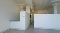 This single-floor space blends industrial character with a clean, contemporary aesthetic. Light hardwood-style flooring runs throughout, lending a cohesive and polished look to the interior. The open layout is defined by crisp white walls and partial partition walls that create natural divisions within the space without fully closing off areas, offering flexibility in how the space can be configured or utilized.

The ceiling is a notable architectural feature, showcasing exposed beams, structural elements, and what appears to be a decorative tin or metallic ceiling finish in portions of the space, adding industrial charm and visual texture overhead. Track lighting is installed along the ceiling, providing adjustable, practical illumination throughout.

A bank of three large windows fitted with vertical blinds allows generous natural light to fill the space while offering views of the surrounding urban environment. The windows also feature a low ledge beneath them, adding a subtle architectural detail.

Overall, the space carries a bright, neutral, and versatile ambiance, well-suited for a variety of uses. The combination of raw industrial elements and modern finishes creates an engaging and functional environment.