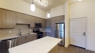 This single-floor space showcases a thoughtfully designed kitchen as its centerpiece. The kitchen features soaring ceilings that create an open, airy atmosphere, enhanced by elegant pendant lighting suspended above the expansive granite-topped island, which provides generous prep and gathering space. A coordinating track lighting system further illuminates the area.

The cabinetry throughout is finished in a warm, textured wood-tone laminate, offering ample storage while contributing to the modern aesthetic. A geometric-patterned tile backsplash adds visual interest behind the countertops. The kitchen is equipped with a full suite of stainless steel appliances, including a gas range, built-in microwave, and a full-size refrigerator, alongside a stainless steel sink.

Hardwood-style flooring runs throughout, adding warmth and continuity to the space. The neutral color palette of soft whites and warm wood tones creates a cohesive and inviting environment. A paneled door is visible, suggesting access to additional areas of the home. Overall, the space balances practicality with refined, contemporary design details.