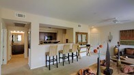 This single-story space showcases an open-concept layout that seamlessly connects the kitchen, dining, and living areas. The kitchen is partially enclosed and visible through a pass-through opening, which doubles as a breakfast bar. Dark-toned cabinetry with granite or stone countertops lines the kitchen, and black appliances, including a microwave and range, are neatly integrated into the cabinetry. The pass-through bar is accompanied by three woven rattan barstools, adding a natural, textural element to the space.

Adjacent to the kitchen bar, a rustic wooden console table with an X-shaped base serves as a decorative accent, topped with a framed mirror and small decorative objects. The flooring throughout the main living area consists of wall-to-wall carpeting in a neutral tone, contributing to a warm and comfortable ambiance. Recessed lighting and ceiling-mounted vents are visible, ensuring consistent climate control. The entryway, visible in the background, features a paneled door with clean white trim, reinforcing the space's bright and cohesive aesthetic. Overall, the layout is functional and well-organized, with a consistent warm, neutral color palette.