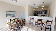 This single-floor residence offers a thoughtfully designed open-concept layout that seamlessly connects the kitchen, dining, and living areas. The kitchen is equipped with dark espresso cabinetry, granite-style countertops, black appliances including a refrigerator, electric range, and built-in microwave, and durable hardwood-style flooring. A raised breakfast bar with pendant lighting provides additional seating and serves as a natural divider between the kitchen and dining space, where a full dining table comfortably accommodates multiple guests.

The living area features plush carpeting and neutral walls that create a warm, versatile backdrop. Two well-proportioned bedrooms are bathed in natural light from large windows with exterior views, and both feature soft carpet flooring. The primary bathroom offers a dual-sink vanity with granite-style countertops, stylish cabinetry, and direct access to a walk-in closet with built-in shelving. A secondary bathroom includes a tub-shower combination with clean white fixtures. Throughout the residence, consistent neutral tones, flush-mount and pendant lighting, and quality finishes contribute to a cohesive, comfortable ambiance.