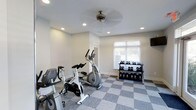 The Aspenwood Apartments fitness center is a dedicated, single-floor workout facility designed to support residents' health and wellness goals. The space features a clean, modern aesthetic with crisp white walls and a stylish checkered gray floor that adds visual interest while providing a durable, easy-to-maintain surface ideal for a gym environment. Recessed ceiling lights ensure consistent, even illumination throughout the room, while a ceiling fan promotes air circulation during workouts. A window fitted with white blinds allows natural light to filter in, creating a bright and welcoming atmosphere. The facility is equipped with a selection of cardio machines, including a spinning bike and an elliptical trainer, offering residents versatile options for cardiovascular exercise. A wall-mounted screen provides entertainment or guided workout content. The free weight area features a multi-tier dumbbell rack stocked with a range of weights, accommodating both beginner and experienced exercisers. The layout is thoughtfully organized to allow comfortable movement between equipment. Overall, the Aspenwood fitness center provides a functional, well-maintained amenity space that meets a broad range of fitness needs in a comfortable and accessible setting.