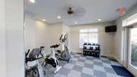 The Aspenwood Apartments fitness center is a dedicated, single-floor workout facility designed to support residents' health and wellness goals. The space features a clean, modern aesthetic with crisp white walls and a stylish checkered gray floor that adds visual interest while providing a durable, easy-to-maintain surface ideal for a gym environment. Recessed ceiling lights ensure consistent, even illumination throughout the room, while a ceiling fan promotes air circulation during workouts. A window fitted with white blinds allows natural light to filter in, creating a bright and welcoming atmosphere. The facility is equipped with a selection of cardio machines, including a spinning bike and an elliptical trainer, offering residents versatile options for cardiovascular exercise. A wall-mounted screen provides entertainment or guided workout content. The free weight area features a multi-tier dumbbell rack stocked with a range of weights, accommodating both beginner and experienced exercisers. The layout is thoughtfully organized to allow comfortable movement between equipment. Overall, the Aspenwood fitness center provides a functional, well-maintained amenity space that meets a broad range of fitness needs in a comfortable and accessible setting.