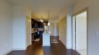 This single-floor residence offers a well-designed open-concept layout that seamlessly connects the kitchen and living areas. The kitchen is a standout feature, showcasing dark espresso cabinetry that contrasts beautifully with a light-toned granite kitchen island, which serves as both a prep space and a casual dining spot. Pendant lighting hangs above the island, adding a modern touch while providing focused illumination. Stainless steel appliances, including a refrigerator, microwave, and dishwasher, complement the sleek mosaic tile backsplash that runs along the cooking wall. Rich, dark hardwood flooring flows throughout the open area, lending warmth and continuity to the space. Striped accent rugs frame the kitchen zone, adding texture underfoot. The layout includes multiple doorways leading to additional rooms, suggesting a practical and well-organized floor plan with generous access to private spaces. Crisp white walls and trim throughout keep the overall aesthetic clean and bright. The combination of dark cabinetry, modern fixtures, and quality flooring gives this residence a refined yet inviting character that balances style with everyday functionality.