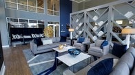 This single-floor space makes a striking impression through its double-height ceilings and expansive upper-level windows, which flood the interior with natural light. The design palette centers on a refined combination of navy blue and cool grey, creating a cohesive and upscale aesthetic throughout.

The main seating area is anchored by plush grey sofas and armchairs accented with deep navy cushions, arranged around a pair of sleek white marble-topped coffee tables. A geometric white lattice shelving unit serves as a bold architectural divider, adding visual depth while displaying curated white décor objects. The space is further elevated by standout accent pieces, including a sculptural gold sunburst ornament, a deep blue glass bowl, and a classic table lamp that contribute warmth and contrast against the cool tones.

Underfoot, a large area rug with bold navy and white geometric patterning defines the lounge zone, while large ground-floor windows and glass entry doors reinforce the connection to the exterior. The overall ambiance is contemporary and professionally curated, suited for a communal or reception-style environment.