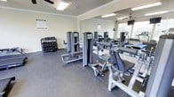 This fitness center offers a focused and functional workout environment across a single floor, thoughtfully designed to accommodate a range of exercise routines. The space features durable rubber flooring throughout, providing both safety and comfort during training sessions. Crown molding details along the upper walls lend a polished, finished look to the room.

The strength training area is well-stocked, with a full rack of dumbbells available in a variety of weights, catering to users of all fitness levels. Multiple cable and resistance machines are positioned throughout the space, offering options for targeted muscle group training. An adjustable bench is also present, adding versatility to free-weight workouts.

Large floor-to-ceiling mirrors line one wall, creating a sense of expanded space while allowing users to monitor their form during exercise. Natural light filters in through a window, complementing the overhead fluorescent lighting and ceiling fan that help maintain a comfortable training environment. A posted fitness center rules notice reflects an organized and community-oriented atmosphere. Overall, this gym provides a comprehensive and accessible space for residents or members seeking a reliable, on-site fitness facility.