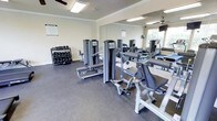 This fitness center offers a focused and functional workout environment across a single floor, thoughtfully designed to accommodate a range of exercise routines. The space features durable rubber flooring throughout, providing both safety and comfort during training sessions. Crown molding details along the upper walls lend a polished, finished look to the room.

The strength training area is well-stocked, with a full rack of dumbbells available in a variety of weights, catering to users of all fitness levels. Multiple cable and resistance machines are positioned throughout the space, offering options for targeted muscle group training. An adjustable bench is also present, adding versatility to free-weight workouts.

Large floor-to-ceiling mirrors line one wall, creating a sense of expanded space while allowing users to monitor their form during exercise. Natural light filters in through a window, complementing the overhead fluorescent lighting and ceiling fan that help maintain a comfortable training environment. A posted fitness center rules notice reflects an organized and community-oriented atmosphere. Overall, this gym provides a comprehensive and accessible space for residents or members seeking a reliable, on-site fitness facility.