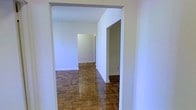 This single-floor space presents a straightforward and functional layout characterized by its most notable feature: richly patterned parquet hardwood flooring that extends throughout the visible areas. The warm, geometric wood blocks create a classic aesthetic that anchors the otherwise clean and minimal interior.

The space is organized with an open corridor-style flow, where a wide, squared archway leads into a main room, allowing for a natural sense of connected spaces. The white walls are smooth and neutral throughout, providing a blank canvas adaptable to various uses. A doorway visible to the right suggests an additional adjoining room or hallway, contributing to a multi-room layout on the single floor.

The ceilings are of standard height, and the baseboard trim runs consistently along the walls, offering a modest but tidy finishing detail. The absence of furniture in the visible areas highlights the clean lines of the architecture and the condition of the flooring. Overall, the space conveys a modest, utilitarian character with the warmth of traditional hardwood underfoot, making it a practical and adaptable environment.