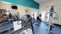 This fitness center is designed to support a well-rounded cardio workout routine within a compact, efficiently arranged layout. The room features rubber flooring throughout, providing a durable and slip-resistant surface suited for exercise equipment and active use. A bold teal accent band runs along the upper portion of the walls, adding a vibrant pop of color against the neutral beige backdrop and giving the space a modern, energetic feel.

Large floor-to-ceiling mirrors line one wall, visually expanding the room and allowing users to monitor their form during workouts. The equipment selection includes treadmills, a stationary recumbent bike, an elliptical-style machine, and additional cardio apparatus, offering variety for different fitness preferences. A wall-mounted flat-screen television is positioned for easy viewing during exercise sessions.

Natural light enters generously through a set of glass-paned French doors, which also provide a view of the surrounding greenery outside. The doors contribute to an open, airy atmosphere within the space. An emergency exit sign and ventilation units are also present, reflecting attention to safety and climate comfort throughout the fitness area.