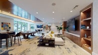 This elegantly designed single-floor amenity space offers a seamless blend of modern aesthetics and functional social areas. The interior is anchored by a striking entryway featuring a warm wood-paneled tray ceiling with ambient cove lighting, glass railings, and a cascading cluster of woven globe pendant lights that serve as an eye-catching focal point.

The main lounge area is expansive and open, highlighted by a bold, artistically patterned area rug that anchors the space beneath multiple seating configurations. Plush sofas and accent chairs in neutral tones with pops of yellow and blue create inviting conversation areas, while a wall-mounted flat-screen TV and built-in wood media wall anchor one end of the room. Large floor-to-ceiling glass doors and windows throughout flood the interior with abundant natural light and provide views of lush outdoor greenery.

A billiards table with bar-height seating offers a dedicated entertainment zone, while a sleek bar counter with additional high stools provides a social gathering spot. Colorful contemporary artwork is displayed throughout, adding vibrancy and personality to the neutral white walls and textured wallcoverings. Recessed lighting and polished tile flooring complete the refined, resort-inspired atmosphere of this thoughtfully curated amenity space.