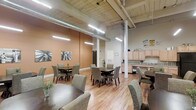 This single-floor space offers an open-concept layout designed for communal dining and gathering. The interior features soaring ceilings with exposed ductwork, structural support columns, and industrial-style pendant lighting that illuminates the room efficiently. The warm terracotta accent walls contrast with the lighter tones of the remaining walls, creating a welcoming ambiance throughout the space.

The main area is furnished with multiple dark-wood dining tables, each surrounded by upholstered grey chairs, providing ample seating for groups. Light-toned hardwood flooring runs throughout, adding warmth and continuity to the industrial aesthetic. Black-and-white artwork adorns the accent walls, contributing a subtle decorative element.

Along one side of the room, a built-in kitchenette features light wood cabinetry, a sink, and counter space, making the area well-suited for food preparation and service. A doorway near the back provides additional access to adjacent areas. The overall layout is practical and spacious, accommodating a variety of communal activities while maintaining a comfortable and inviting atmosphere.