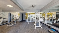 Located at 1160 N Conwell Ave in Covina, California, this fitness center offers a well-appointed, single-floor space designed to support a comprehensive workout experience. The room is finished with durable, multi-tone carpet flooring and features a textured ceiling with a ceiling fan that promotes air circulation throughout. Bold yellow accent walls paired with a striking geometric white feature wall give the space a modern, energetic aesthetic, while large windows and glass doors flood the interior with natural light and provide direct views of an outdoor pool area and surrounding landscaping. Full-length mirrors line one wall, visually expanding the space and allowing users to monitor their form during exercise. The fitness center is equipped with a wide range of Matrix-brand cardio equipment, including treadmills, elliptical trainers, and stationary bikes, as well as multi-station cable and strength training machines. The thoughtful layout separates cardio and strength training zones, enabling comfortable use by multiple individuals simultaneously. Recessed and flush-mounted ceiling lights supplement natural lighting for evening use. Overall, the space delivers a clean, functional, and motivating environment well-suited for residents seeking convenient, on-site fitness amenities in the Covina area.