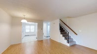 Located at 200F Summitwood, this multi-level home spans four floors, offering a generous and well-organized living arrangement across its various levels. The main level showcases light hardwood-style flooring that flows seamlessly through the open living area, creating a warm and inviting atmosphere. A central staircase with a dark metal railing and wooden handrail serves as a functional focal point, connecting the floors with an understated elegance. The open layout allows for a natural flow between the living area and the adjacent kitchen, which is visible through a rounded archway. The kitchen is finished with white cabinetry and benefits from natural light streaming in through a bright window. A pendant light fixture adds a classic touch above the kitchen space. Neutral white walls throughout contribute to a clean, airy aesthetic, while baseboard heating units indicate practical, year-round comfort. Additional doorways hint at further rooms and utility spaces beyond the main living area. The overall ambiance is bright, functional, and move-in ready, making this four-floor home a versatile space suitable for a variety of living arrangements.