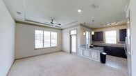 This single-floor residence offers 2 bedrooms and 2 bathrooms within an open-concept layout that connects the living area and kitchen. The living room features plush carpeting, a tray ceiling with crown molding, and a ceiling fan, adding both elegance and comfort. Large windows fitted with horizontal blinds allow generous natural light to fill the space, creating a bright and welcoming atmosphere.

The kitchen is positioned adjacent to the living area and is defined by dark cabinetry, granite or stone-look countertops, and pendant lighting hanging above a peninsula-style breakfast bar. The bar provides additional workspace and casual seating. Track lighting above the kitchen adds further illumination, complementing the pendant fixtures.

The transition between the kitchen and living room is seamless, making the layout well-suited for everyday living and entertaining. The neutral color palette throughout — warm beige walls, light carpeting, and dark cabinetry accents — creates a cohesive and balanced aesthetic. Crown molding details throughout the space enhance the overall polished and refined character of this residence.