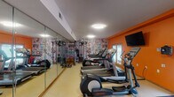 Located at 3560 Minikahda Ct in St. Louis Park, Minnesota, this fitness center is a purpose-built exercise facility designed to support a well-rounded workout experience. The space is contained on a single floor and immediately distinguishable by its bold, energetic interior design — vibrant orange accent walls are paired with a striking comic-style graphic wallcovering that lines one side of the room, creating a dynamic and visually engaging atmosphere.

The main workout area is well-appointed with a lineup of Life Fitness cardio equipment, including multiple treadmills and elliptical cross-trainers arranged in a row. A wall-mounted flat-screen television is positioned for easy viewing during cardio sessions. A cable strength training machine and free weights round out the equipment offerings, providing options for both cardiovascular and resistance training. Full-length mirrors run along one wall, enhancing the sense of space while allowing users to monitor their form.

Natural light filters into the room through windows and glass-paneled doors that look out to an exterior area. Flush-mount ceiling lights provide consistent overhead illumination throughout. An entry corridor with coat hooks, a water cooler, and floor mats offers a practical transition zone into the fitness area. An accessible restroom with grab bars and slate-tile flooring is also available on-site, completing the facility's amenities.