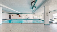 This generously sized single-floor recreational facility offers a diverse range of amenities designed for leisure and activity. The centerpiece of the space is a large indoor swimming pool, surrounded by mosaic-tiled flooring and framed by full-length windows that invite natural light and offer views of the surrounding greenery and waterfront. Teal-accented pipework and structural columns add a distinctive visual character to the pool area's textured white ceiling. A wall of mirrors further enhances the sense of openness, reflecting both the pool and the natural surroundings. Adjacent to the main pool sits a hot tub, accessible via handrail entry steps. A carpeted lounge area along the poolside provides space for relaxation, complete with posted pool rules and safety equipment. The games room features a full-size billiards table with overhead pendant lighting, a dartboard, and bar-height seating, making it an ideal social gathering space with bright fluorescent lighting and light-toned tile flooring. Rounding out the facility is a dedicated squash court with a vivid yellow floor, red boundary lines, and overhead fluorescent lighting — ideal for an active workout. The facility also includes change room access and clearly marked entry and exit points throughout.