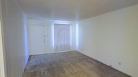 This single-floor space presents a clean and functional layout with a straightforward design. The main living area features wall-to-wall carpeting in a neutral tone, providing a soft and comfortable underfoot feel that complements the room's simple, understated aesthetic. White walls throughout give the space a bright, open atmosphere, while the textured ceiling adds a subtle layer of dimension to the interior. A prominent floor-to-ceiling window fitted with vertical blinds serves as the focal point of the main room, allowing natural light to filter in and illuminate the space. A solid white entry door is positioned adjacent to the window, offering a clear and accessible entryway. The overall ambiance is minimal and move-in ready, with a neutral color palette that provides a versatile backdrop for a variety of furnishing and décor styles. The single-floor layout ensures ease of navigation, making the space practical and straightforward in its use. The combination of natural light, neutral tones, and carpeted flooring creates a comfortable and livable environment suited for everyday use.